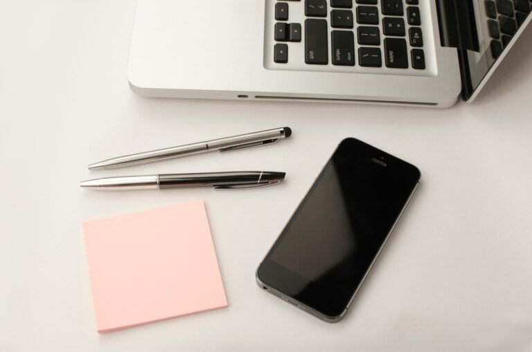 Top is an open laptop with two pens, pad, phone right next to it